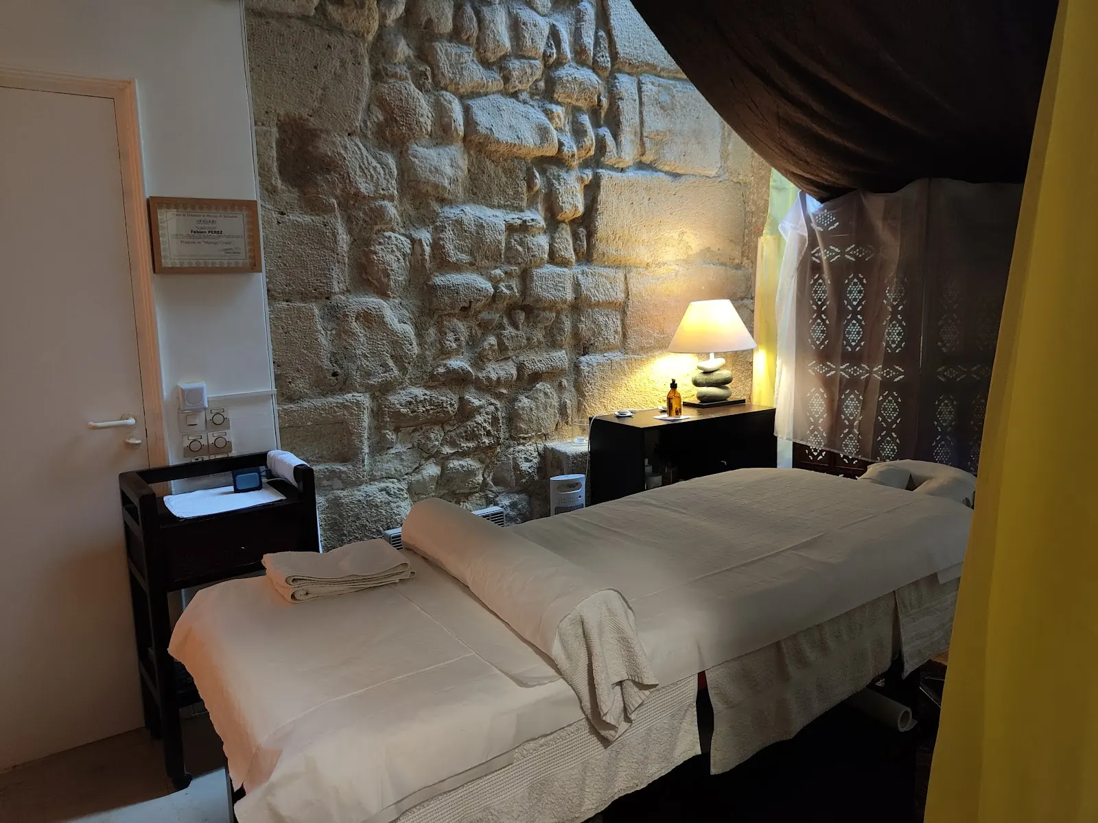 La salle de massage : table au drap blanc, mur de pierre éclairé par une lampe au pied de galets.